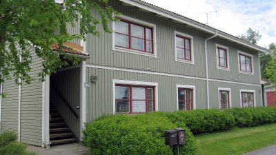 Grönt tvåvånings flerbostadshus i trä. Framför huset är en häck. En trapp vid gaveln av huset som leder upp till andra våningen.