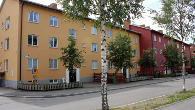 Två lägenhetshus som sitter ihop. Det ena är i gul puts och det andra är rött. Träd planterade utanför entréerna.