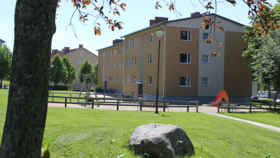 Trevåningshus i lejongul puts. Liten lekplats intill huset och sten och trädstam i förgrunden.