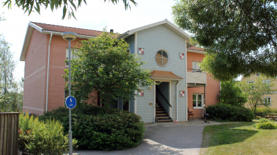 Ljust tegelrött flerbostadshus i två våningar. Grönskande träd och buskar i förgrunden.