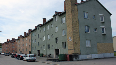 Edelsbergsvägen 17, ett lägenhetshus i puts i tre våningar. Entrédörrar ut mot gatan där det står bilar parkerade.