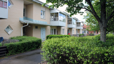 Långt tvåvåningshus i ljusgult tegel. Buskar och träd i förgrunden.