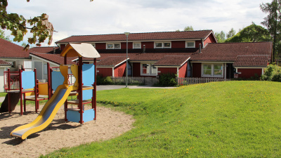 Översiktsbild med grönska och lekplats i förgrunden. I bakgrunden tre röda radhus.