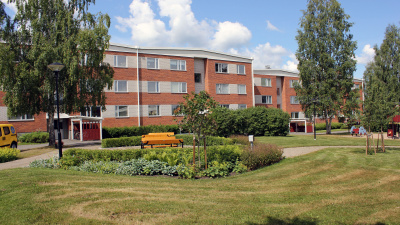 Lägenhetshus i tre våningar i bakgrunden och grönskande rabatt och gräsmatta i förgrunden.