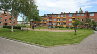 Gräsmatta och lekplats i förgrunden. I bakgrunden två flerbostadshus i tre våningar och rött tegel.