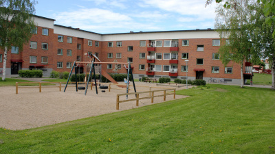 Lekplats i förgrunden och och i bakgrunden ett tegelrött flerbostadshus med röda balkonger i tre våningar.