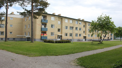 I förgrunden gräsmatta med grusgångar och i bakgrunden gult flerbostadshus i tre våningar.