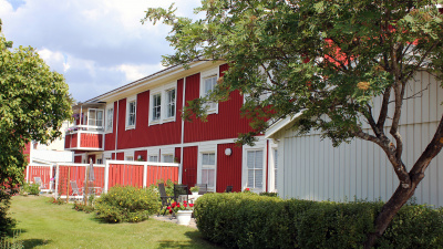 Baksidan av ett flerbostadshus med röd träfasad. Huset är i två våningar och lägenheterna i markplan har uteplats med stenplattor.