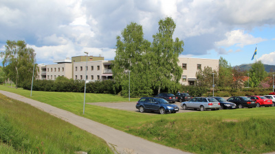 Exteriörbild som på långt håll visar ett stort hus i vitt tegel. I förgrunden en bilparkering och två uppvuxna björkar.