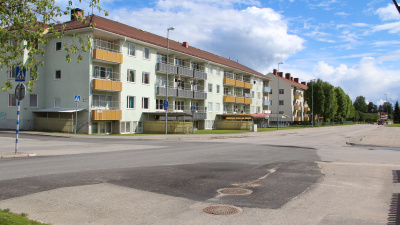 Exteriörbild som visar stort trevåningshus från baksidan med gatukorsning i förgrunden. Huset har källaringångar till hyresgästlokal och bovärdskontor.