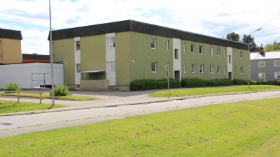 Exteriörbild på flerbostadshus på Skolmästaren i Arbrå. Ett grönt hus i två plan med svart takfot. Bilden visar baksidan med vita fält runt entréerna.