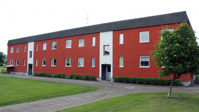Exteriörbild av innergård på Skolmästaren i Arbrå. Bild på ett rött hus i två plan med svart takfot. Bilden visar entrésidan och har vita fält vid ingångarna.
