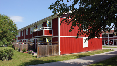 Flerbostadshus i två våningar med röd träfasad. Balkonger på andra våningen och uteplats i markplan.