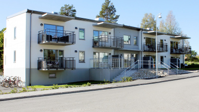 Entrésida av flerbostadshus i två våningar. En stentrapp leder upp till entrédörr. Samtliga lägenheter har svarta balkonger.