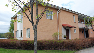 Flerbostadshus i ljust tegel i två våningar. Bilden visar en entrédörr i markplan. Framför huset står ett träd samt buskar.