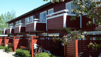 Exteriörbild av entrésida av radhuslägenheter i röd träfasad. Plats för cyklar vid entrén och balkonger på andra våningen.