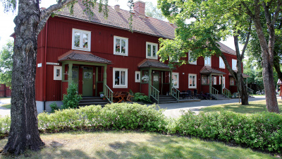 Grönska i förgrunden med gräsmatta och buskar. I bakgrunden ett flerbostadshus i två våningar med röd träfasad. Lägenheterna har egen ingång.