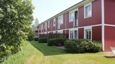 Baksidan av ett flerbostadshus i två våningar. Lägenheter i markplan har uteplats med plattor som omsluts av gröna buskar. Lägenheter på andra våningen har fransk balkong.