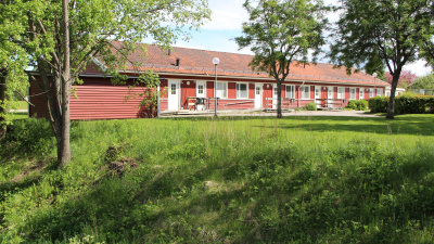 Entrésidan av radhuslänga i ett plan. Huset är rött med rött tegeltak. Gräs och buskage i förgrunden.