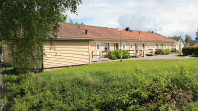 Entrésidan av radhuslänga i ett plan. Huset är gult med rött tegeltak. Gräs och buskage i förgrunden.