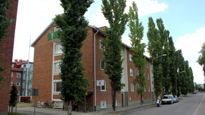 Tegelröda lägenhetshus i tre våningar. Framför huset jäms med gatan står höga smala träd planterade med några meters mellanrum.