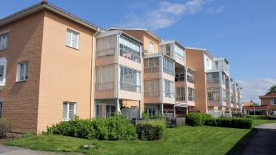 Flerbostadshus i tre våningar med inglasade balkonger och uteplatser i markplan. Gräsmattor och buskar omsluter uteplatser i markplan.