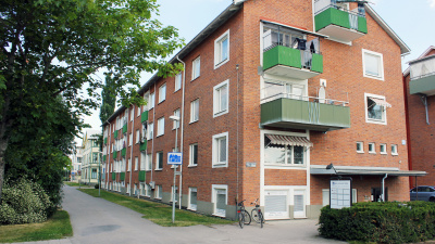 Rött flerbostadshus i tegel och i tre våningar. Bilden är tagen från kortsidan. Till vänster en gång och cykelväg och i botten av huset finns en lokal.