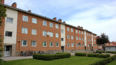 Flerbostadshus i i tre våningar. Rött tegel med ljusa partier vid entréerna. Gröna gräsmattor och buskar i förgrunden.