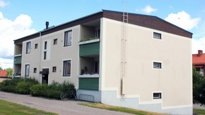 Flerbostadshus i ljus puts. Bilden visar gaveln på huset och man kan se att huset är i suterräng. Lägenheter i markplan har uteplats och lägenheter på första och andra våningen har balkong.