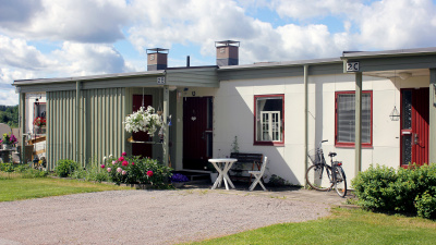 En entré av enplans radhuslänga. Liten trädgårdsmöbel framför entrén. Blommor i krukor och rabatter framför huset. En cykel parkerad framför entrén.