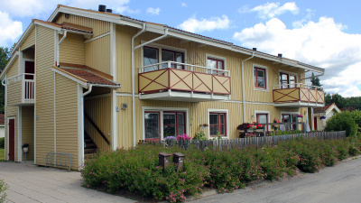 Gult tvåvånings flerbostadshus i trä. Balkonet på andra våningen. En trapp leder till andra våningen. Buskar framför huset.