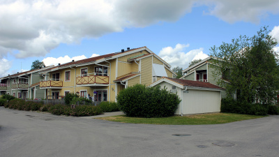 Översikt av bostadsområde med flerbostadshus i två våningar med träfasad. Framför huset en förrådsbyggnad. Lägenheter på andra våningan har balkong.