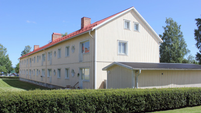 Flerbostadshus i två våningar med gul träfasad. Gröna buskar i förgrunden.